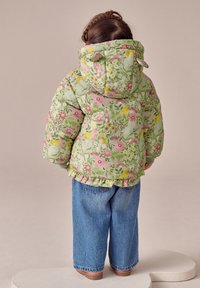 Pamušaluotas žalias žieduotas švarkelis su rožiniais ir geltonais raštais, volanais apatinėje dalyje ir gobtuvu, derinamas su platėjančiomis mėlynomis džinsinėmis kelnėmis.