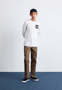 Wit shirt met lange mouwen met een zwart logopatch, bruine broek, zwarte pet en zwarte sneakers met witte zolen, tegen een neutrale achtergrond.