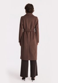 Brązowy płaszcz z paskiem, z wysokim kołnierzem, długimi rękawami i prostą sylwetką. Gładka tekstura, zestawiony z ciemnymi spodniami typu wide-leg.
