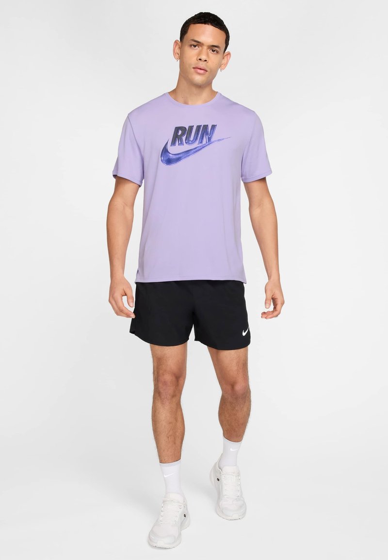 Homme portant un t-shirt Nike violet clair avec l'inscription "RUN", un short Nike noir, des chaussettes blanches et des chaussures de course blanches, debout devant un fond blanc.