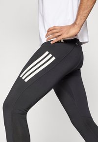Leggings athlétiques noirs avec une texture lisse, comportant trois bandes verticales blanches sur le côté gauche et une coupe ajustée.
