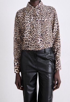 Osoba ubrana w krótki, zapinany na guziki top w cętki lamparta oraz wysokie, czarne skórzane spodnie z bocznymi kieszeniami, stojąca na tle jednolitego tła.