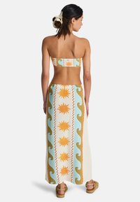 Μπικίνι τοπ χωρίς τιράντες με σχέδιο σε πορτοκαλί, τιρκουάζ και λευκό; μακρύς φούστα με μοτίβα ήλιου και κυμάτων; εμφανή πλεγμένα σανδάλια.