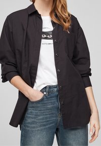 Svart skjorta med knappar och krage, avslappnad passform och långa ärmar, bärs över en vit grafisk t-shirt och blå jeans.