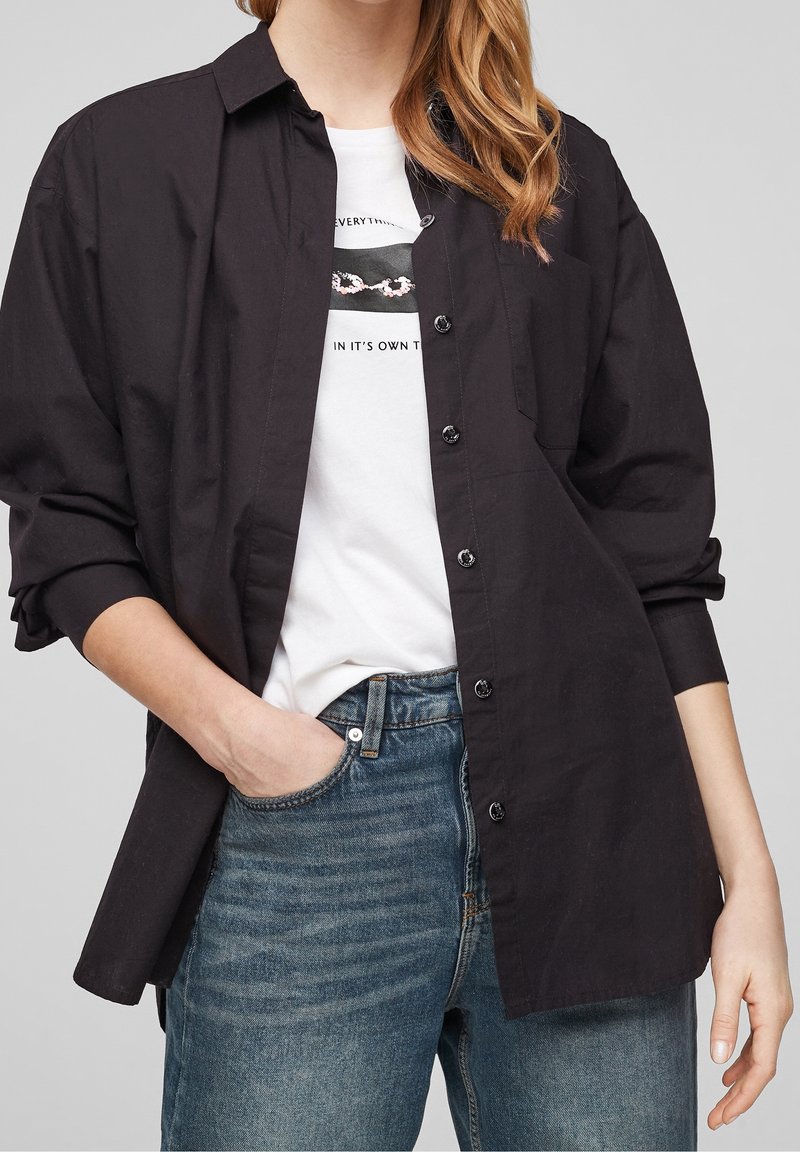 Svart skjorta med knappar och krage, avslappnad passform och långa ärmar, bärs över en vit grafisk t-shirt och blå jeans.