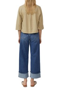 Khaki cropped shirt met 3/4 mouwen gecombineerd met losse blauwe denim jeans, met omgeslagen zoom en achterzakken. Casual schoenen zichtbaar.