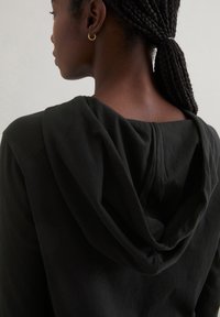 Zwarte hoodie met een relaxte pasvorm, zachte stof en een gevoerde capuchon. Beschikt over subtiele stiksels en een plooidetail aan de achterkant van de halslijn.