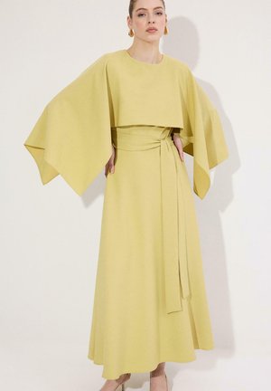 Γυναίκα με μακρύ, ανοιχτό κίτρινο φόρεμα με φαρδιά, ρέοντα μανίκια και δεμένη ζώνη στη μέση, που στέκεται μπροστά σε απλό λευκό φόντο.