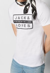 White cotton t-shirt featuring a black rectangular graphic with "JACK & JONES" text and additional branding. Pairing with light blue jeans.