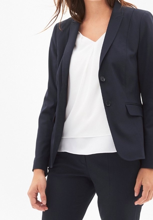 Femme portant un blazer bleu marine et un pantalon avec un chemisier blanc à col en V, debout devant un fond blanc uni.