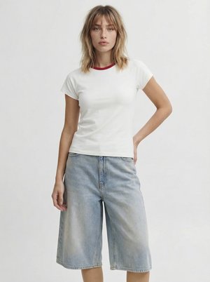 Femme aux cheveux mi-longs portant un t-shirt blanc moulant avec un col rouge et un short long en jean bleu clair, debout devant un fond uni.