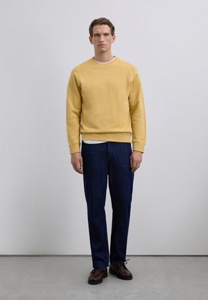 Man staat tegen een witte muur gekleed in een gele sweatshirt, wit hemd, donkerblauwe jeans en bruine leren schoenen.