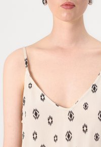 Vaaleanbeige ilmanhiha-toppi, jossa on V-muotoinen kaula-aukko, mustilla geometrisilla kuvioilla ja ohuilla olkaimilla, valmistettu pehmeästä, teksturoidusta kankaasta.
