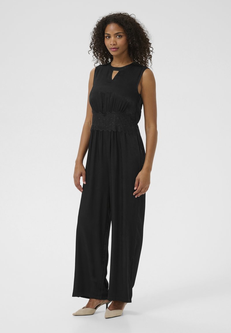 Combinaison noire avec un décolleté en trou de serrure, détail en dentelle à la taille et design à jambes larges. Le tissu lisse et le corsage ajusté mettent en valeur la silhouette.