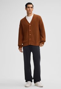 Cardigan lavorato a maglia marrone con scollo a V, dotato di bottoni grandi e polsini a coste, abbinato a pantaloni neri a gamba larga e sneakers bianche.