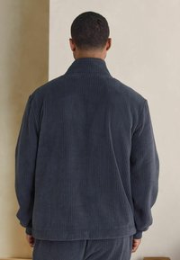 Dark gray corduroy jacket with a high collar, ribbed texture, and relaxed fit, featuring subtle seams and matching pants visible at the bottom.