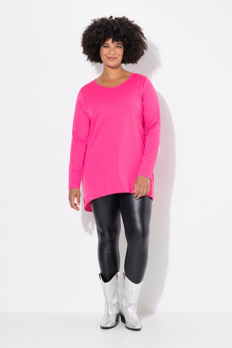Donna con capelli ricci che indossa una tunica a maniche lunghe rosa acceso, leggings in pelle neri e stivali da cowboy argentati, in piedi su sfondo bianco.