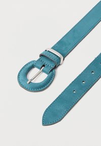 Sininen mokkanahkavyö, jossa on pyöreä hopeinen solki ja useita säätöreikiä valkoista taustaa vasten.