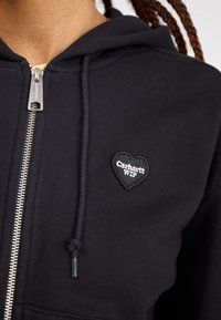 Felpa nera con zip realizzata in misto cotone, dotata di una toppa a forma di cuore con il logo "Carhartt WIP" sul lato sinistro del petto. Dettagli in zip argentata.