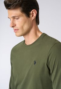 T-shirt a maniche lunghe verde oliva realizzato in tessuto morbido, con un collo rotondo e un piccolo logo blu sul lato sinistro del petto.