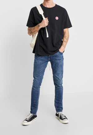 Man in een zwart T-shirt met logo, blauwe jeans, zwarte sneakers en met een witte rugzakstrap over één schouder, staande op een witte vloer.