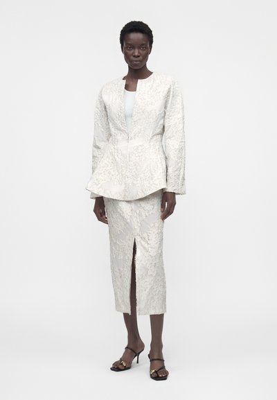 Γυναίκα φοράει υφασμένη λευκή μακρυμάνικη ζακέτα και αντίστοιχη μίντι φούστα με σκίσιμο στο μπροστινό μέρος, συνδυασμένη με μαύρα ανοιχτά πέδιλα με χρυσές λεπτομέρειες.