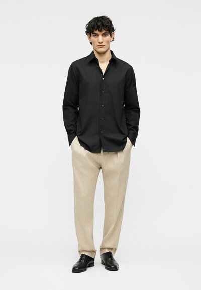 Άντρας με σγουρά σκουρόχρωμα μαλλιά, που φοράει μαύρο πουκάμισο με κουμπιά, μπεζ παντελόνια και μαύρα παπούτσια, στέκεται με τα χέρια στις τσέπες.