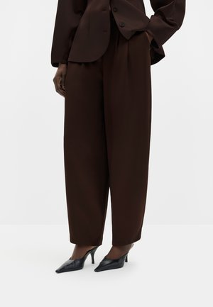Personne portant un pantalon large marron foncé et un blazer assorti, associé à des chaussures à talons aiguilles noires pointues, debout les mains dans les poches.