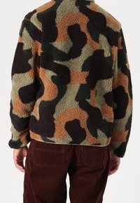 Person som bär en fleecejacka med grön, brun och svart camouflage-mönster, tillsammans med mörkbruna kordfläckbyxor, sedd bakifrån.