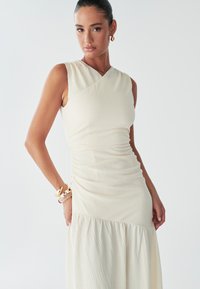 Une femme pose avec confiance dans une robe crème ajustée à la finition texturée, affichant des accessoires minimaux et une coiffure épurée.