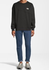 Felpa nera con finitura testurizzata, caratterizzata da un design diagonale e una piccola toppa con logo. Abbinata a jeans in denim blu e sneakers chiare.