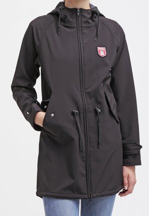 Veste noire à capuche avec fermeture à glissière, cordons de serrage, poches à boutons-pression et un petit patch rouge et blanc sur la poitrine, portée avec un jean bleu.