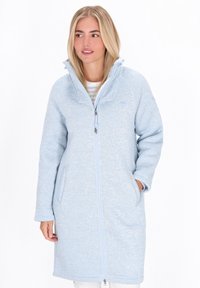 Donna che indossa un cappotto in pile con zip di colore blu chiaro e colletto alto, con le mani nelle tasche e lo sguardo leggermente rivolto verso sinistra.