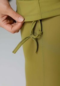 Olijfgroene kledingstuk met een zachte textuur. Bevat een trekkoord met een geknoopt detail en zichtbare stiksels op de zoom.