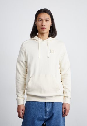 Cream hoodie with a front pocket, drawstring hood, ribbed cuffs, and a small logo patch on the chest, paired with blue denim pants.