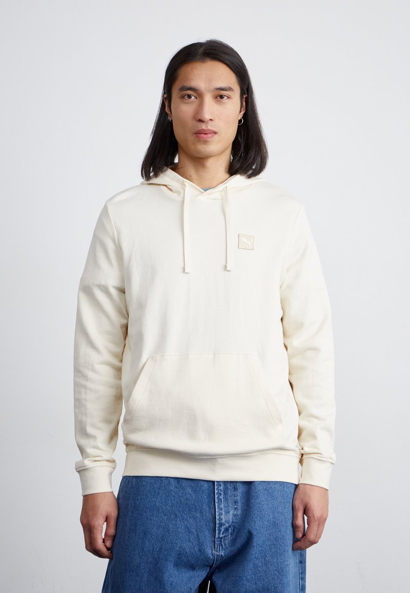 Cream hoodie with a front pocket, drawstring hood, ribbed cuffs, and a small logo patch on the chest, paired with blue denim pants.