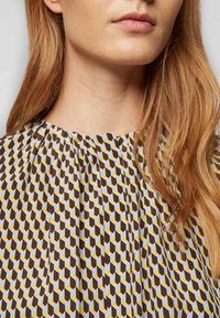 Top à col halter présentant un motif géométrique dans des tons de marron, jaune et bleu. Le tissu a une texture douce avec des plis le long de l'encolure.