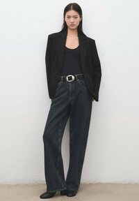 Blazer negro a medida, top negro ajustado, pantalones anchos de mezclilla oscura de talle alto, hebilla de cinturón plateada y zapatos negros sin cordones contra una pared clara.