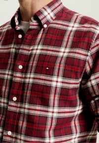 Homme portant une chemise à carreaux rouge, blanche et noire boutonnée sur un T-shirt blanc, laissant apparaître le col et la partie supérieure de la poitrine.