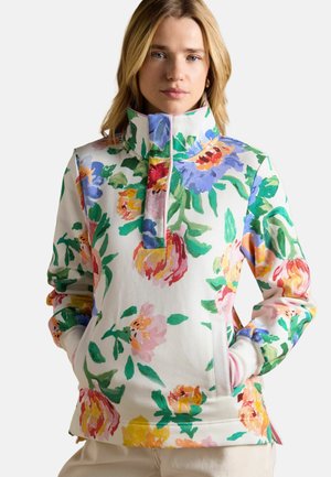 Woman wearing a high-collar pullover with large multicolored floral print, hands in front pockets, neutral background.