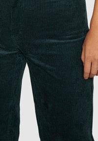 Mörkgröna kordbyxor med vertikalt ribbad textur. Nära bild som visar tygets glans och en hand som vilar på lårområdet.