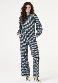 Geprinte twee-delige outfit met geometrische driehoeken in zwart, wit en blauw. Lange mouwen, losse pasvorm en rechte broekspijpen. Metalen schoenen.