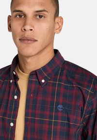Camisa de cuadros en rojo oscuro y azul marino con acentos amarillos, con cuello botón y logo bordado en el pecho.
