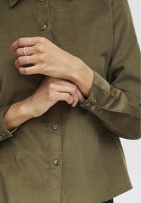 Camicia in corduroy verde oliva con colletto, chiusura con bottoni e maniche lunghe. Presenta bottoni marroni e una vestibilità leggermente cropped.