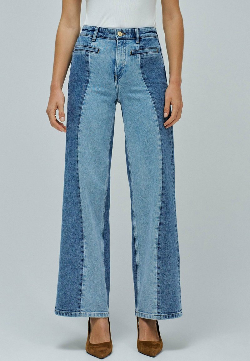 Jeans de cintura alta y pierna ancha con paneles de denim contrastantes en azul claro y oscuro, bolsillos frontales y tacones de punta marrones.