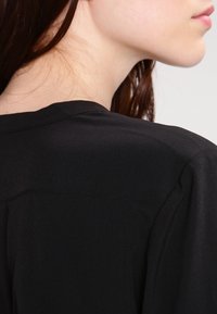 Schwarze Bluse aus glattem, leichtem Stoff, mit einem einfachen Kragen und dezenten Nahtdetails auf der Rückseite.