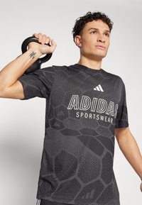 T-shirt atlético preto com uma superfície texturada e padrão, logótipo "ADIDAS" em branco, mangas curtas, emparelhado com um kettlebell preto.