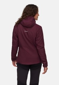 Vrouw die wegkijkt en een maroon Mammut-hoodie en zwarte broek draagt, voor een neutrale achtergrond.