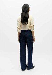 Tmavě modré džínové kalhoty s širokými nohavicemi a vysokým pasem, doplněné světlým krémovým svetrem. Významné detaily zahrnují zadní kapsy a čisté zakončení.