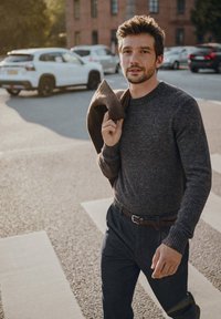 Szary sweter dzianinowy, ciemne spodnie, trzyma brązowy płaszcz. Tekstura wydaje się miękka; sweter ma prążkowane mankiety. Luźne, miejskie otoczenie na zewnątrz.